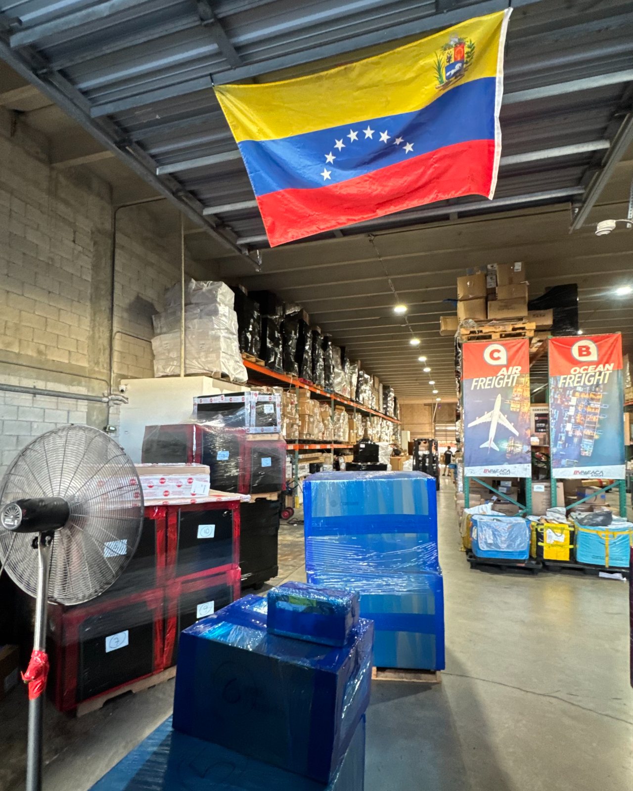 The warehouse floor at BVA & EACA Export in Miami: shrink-wrapped pallets, the racks, and the Venezuelan flag over the aisle